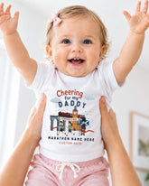 A smiling baby girl wearing a white t-shirt with a graphic design and text, being held up by someone's arms.