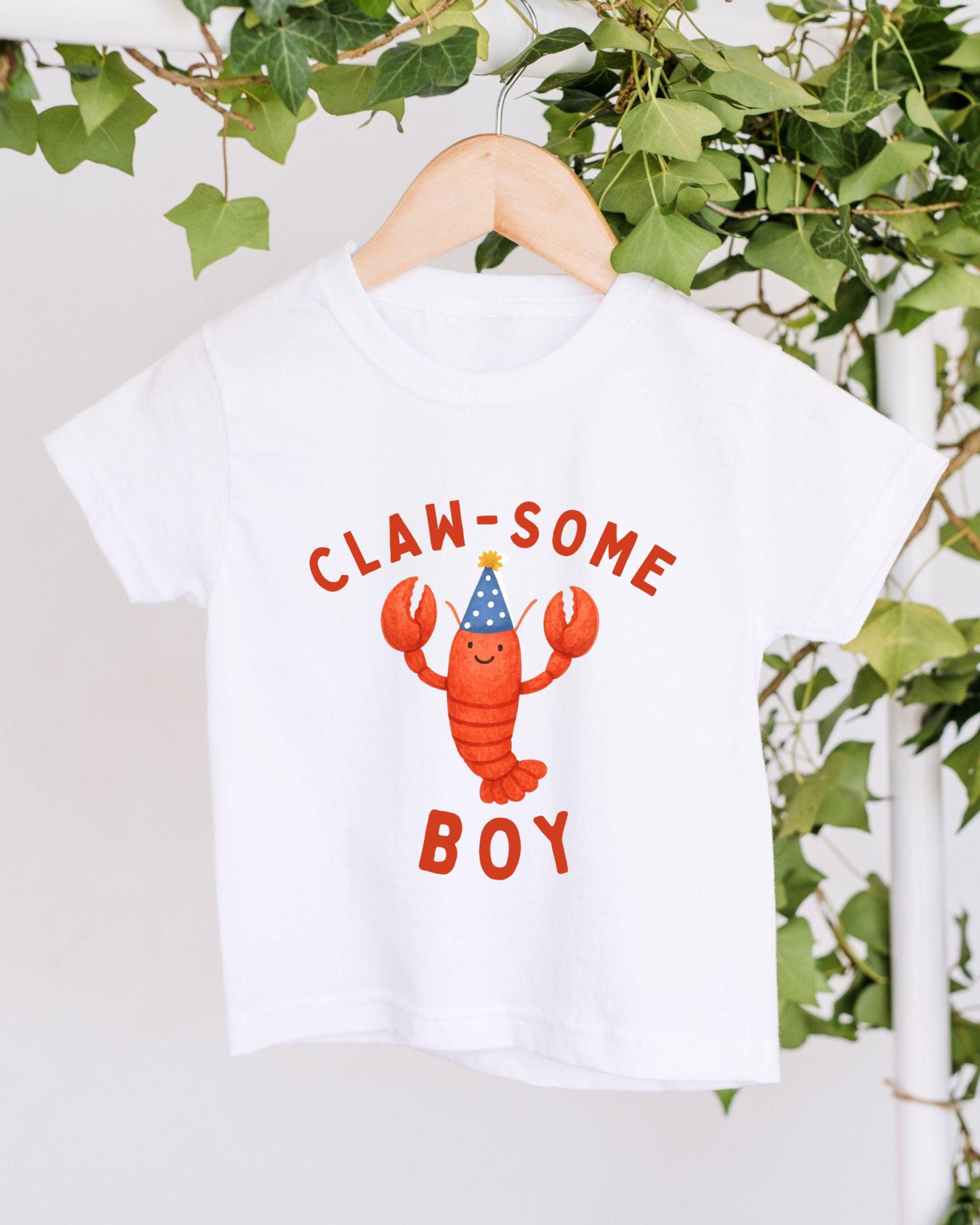 A white t-shirt with a lobster wearing a party hat and the text "CLAW SOME BOY" printed on it.