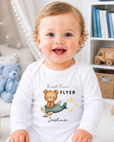 A smiling baby boy wearing a white onesie with a teddy bear and airplane design, sitting in a room with bookshelves in the background.