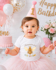 A young girl wearing a white t-shirt with a bee design and a pink birthday hat sits in front of a birthday cake and balloons, with a "Happy Birthday" sign in the background.