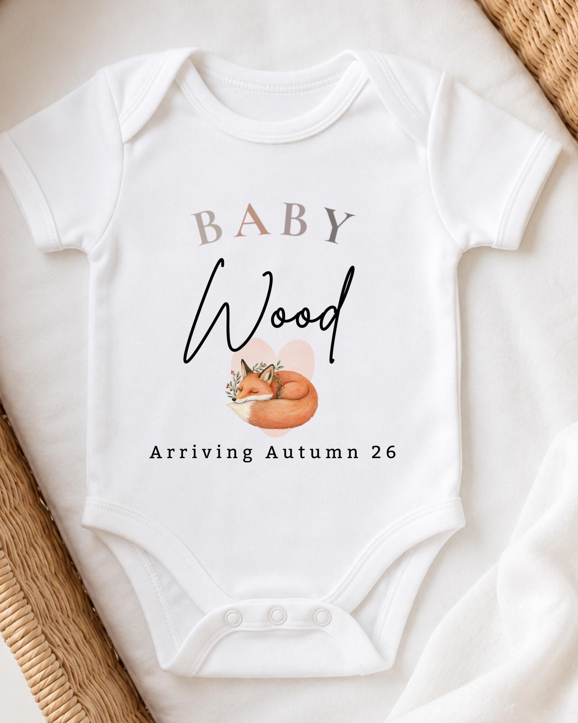 A white baby onesie with the text "BABY Wood Arriving Autumn 26" printed on it, along with an illustration of a fox.