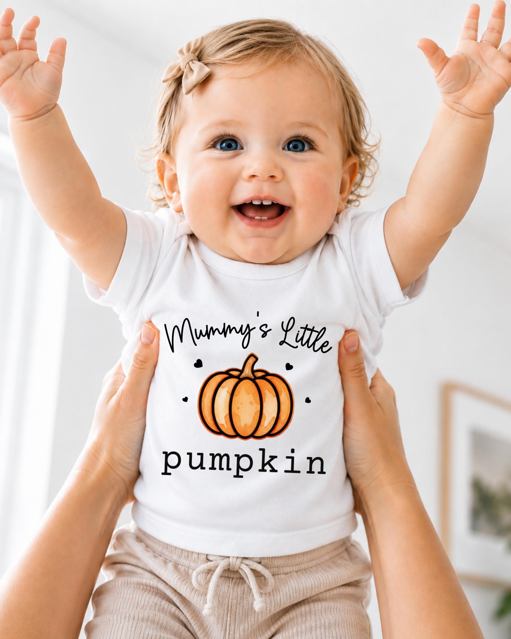 A smiling baby wearing a white t-shirt with a pumpkin graphic and the text "Mummy's Little pumpkin" being held up by two hands.