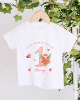 A white t-shirt with a personalized design hanging on a wooden hanger, featuring a fairy character and the text "When I wake up I will be 1 Amy".