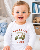 A smiling baby wearing a white onesie with a golf cart graphic and the text "NEW TO THE CLUB" printed on it.