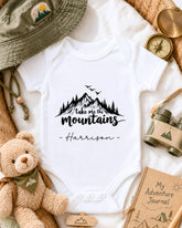 A baby onesie with the text "Take me the mountains" printed on it, surrounded by various outdoor-themed items such as a hat, compass, binoculars, and a teddy bear.