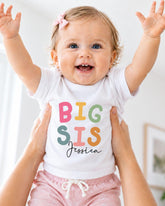 A smiling baby girl wearing a white t-shirt with the text "BIG SIS Jessica" is being held up by someone's arms.
