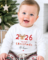A smiling baby boy wearing a white sweater with the text "26 my first christmas Hudson" printed on it, sitting in front of a Christmas tree.