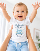A smiling baby wearing a white onesie with a blue car graphic and the text "Halfway to One Scoop" is being held up by someone's arms.