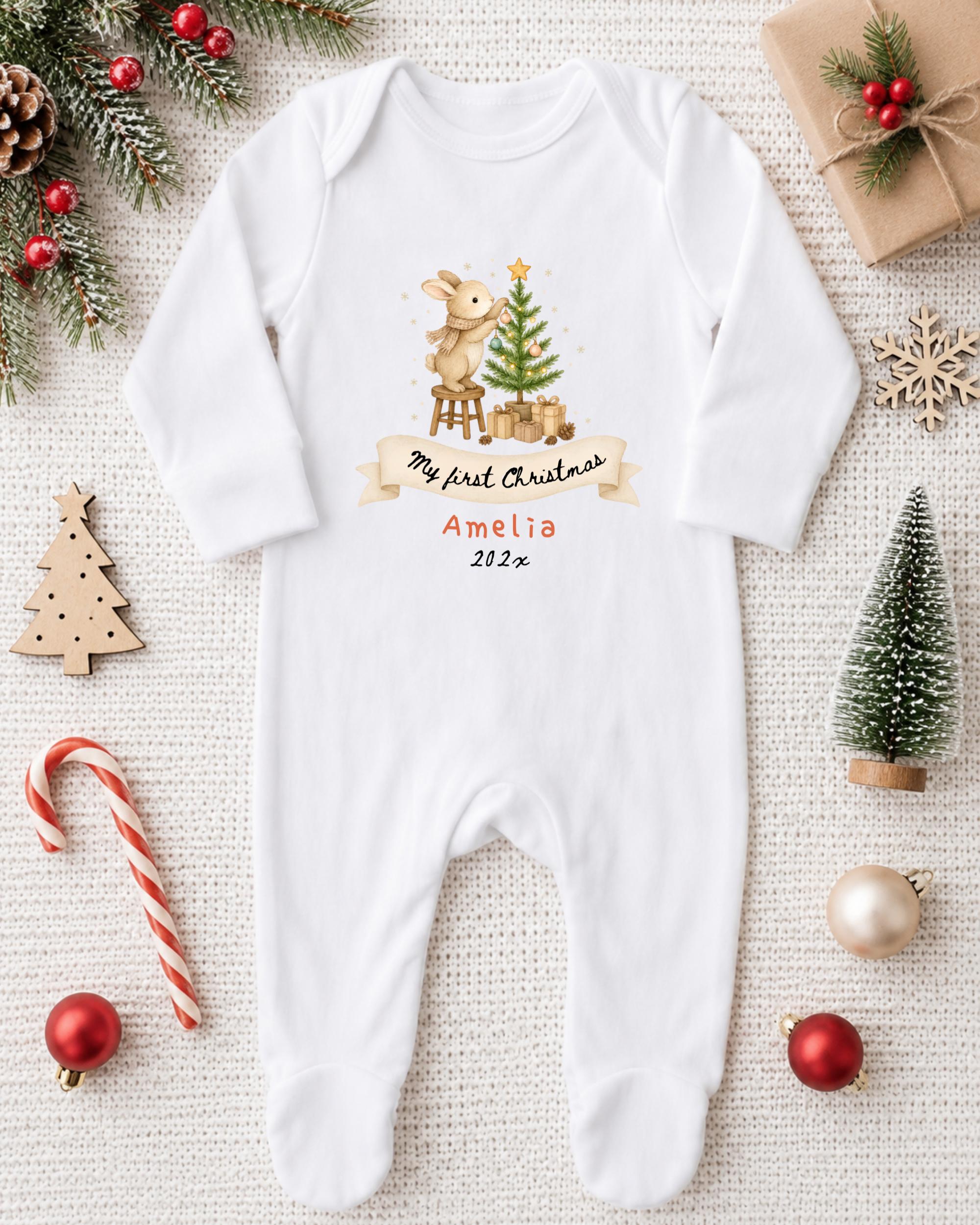 A white onesie with a Christmas-themed design, featuring a bunny and a Christmas tree, is displayed on a white surface alongside various Christmas decorations and gifts.