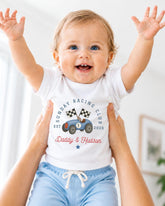 A smiling baby wearing a white t-shirt with a graphic of a race car and the text "Sunday Racing Club EST. Daddy & Hudson" being held up by two people.