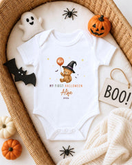 A white onesie with a teddy bear and Halloween-themed decorations, including a ghost, pumpkin, and "BOO!" sign, is displayed in a basket.