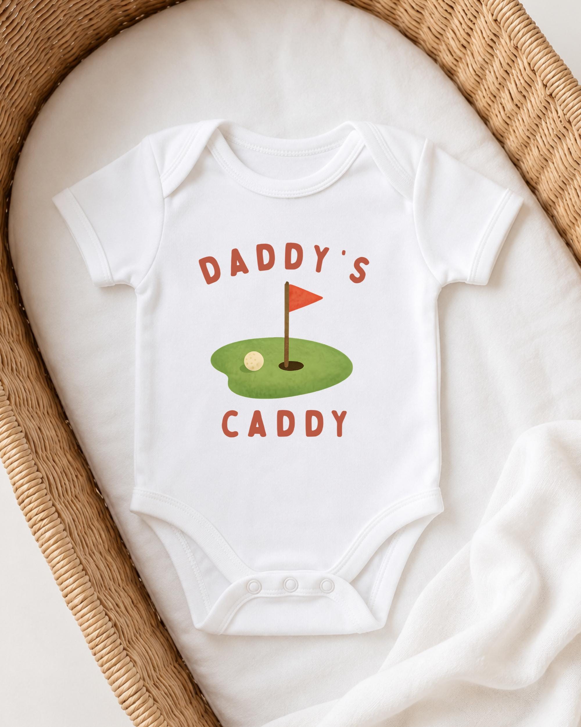 A white onesie with a golf hole graphic and the text "DADDY'S CADDY" printed on it, laid out on a white cloth.