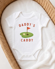 A white onesie with a golf hole graphic and the text "DADDY'S CADDY" printed on it, laid out on a white cloth.