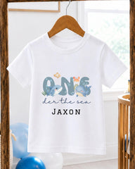 A white t-shirt with a graphic design featuring a cartoon image of a whale and the text "One der the sea JAXON" printed on it.