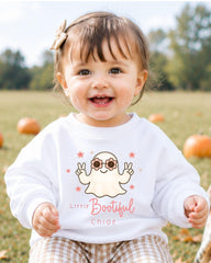 A smiling baby girl wearing a white sweater with a cartoon ghost and the text "Little Bootful Chloe" is sitting in a field with pumpkins in the background.