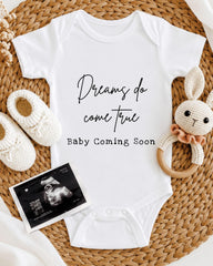 A white baby onesie with the text "Dreams do come true" printed on it, along with a crocheted bunny toy and a baby announcement.