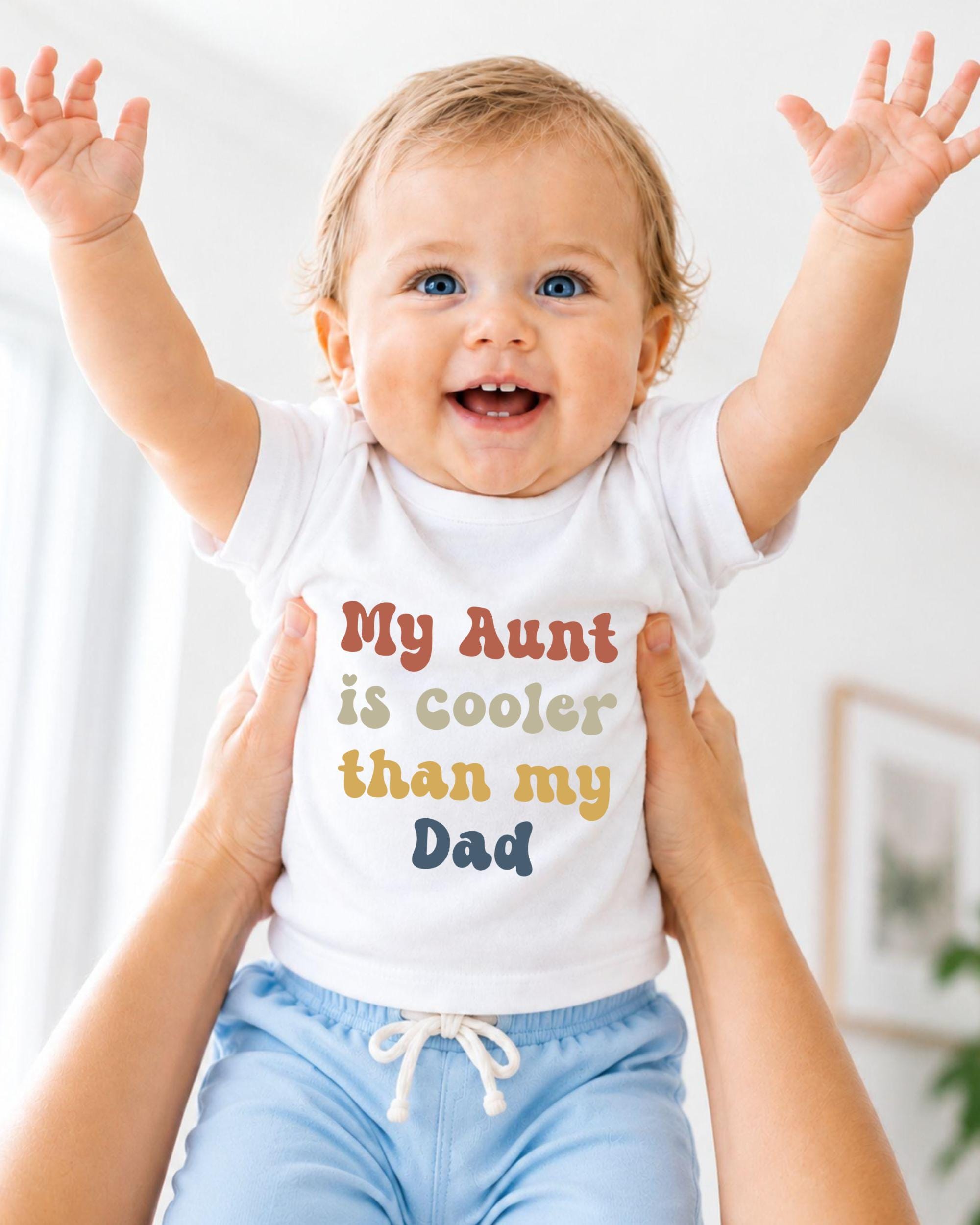 A smiling baby wearing a white shirt with the text "My Aunt is cooler than my Dad" is being held up by someone's arms.