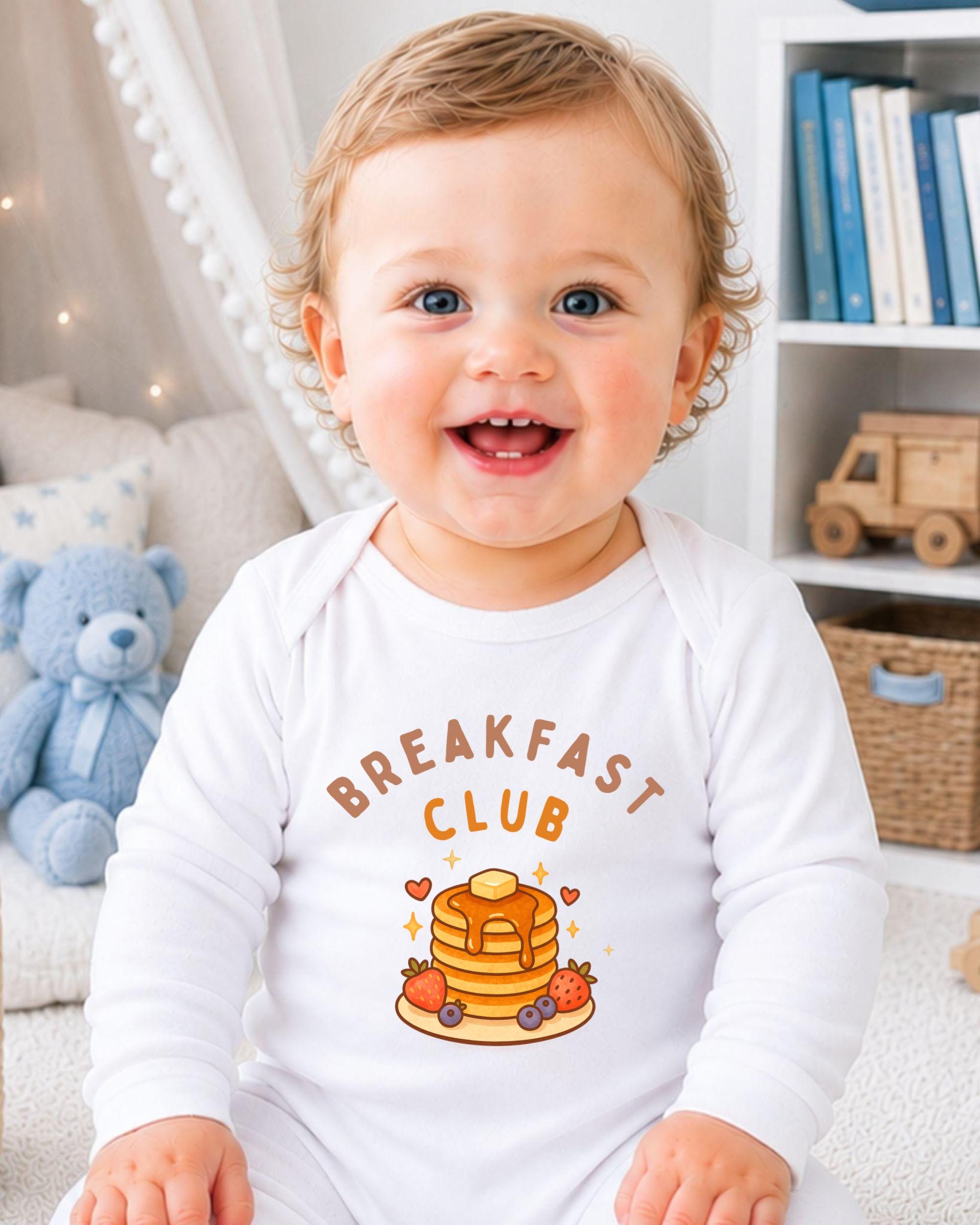 A smiling baby wearing a white onesie with a breakfast club graphic and text.