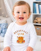 A smiling baby wearing a white onesie with a breakfast club graphic and text.