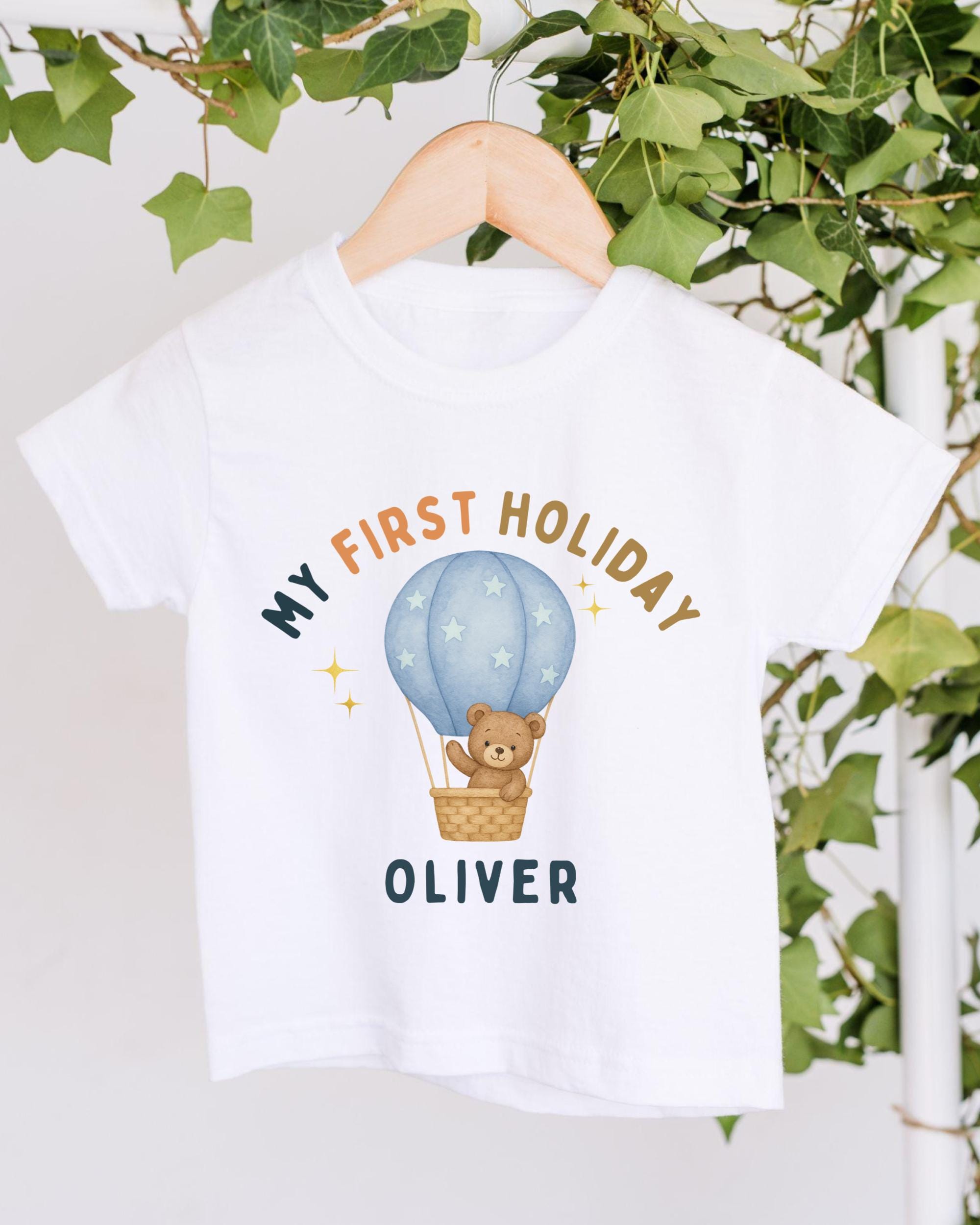 A white t-shirt with a graphic design featuring a teddy bear in a hot air balloon, along with the text "My first holiday Oliver".