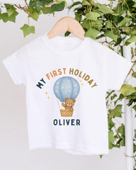 A white t-shirt with a graphic design featuring a teddy bear in a hot air balloon, along with the text "My first holiday Oliver".