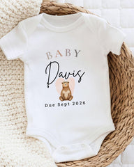 A white baby onesie with the text "BABY Davis Due Sept 2026" printed on it, along with an illustration of a bear.