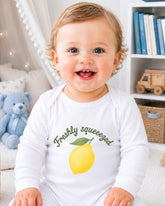 A smiling baby wearing a white onesie with a lemon graphic and the text "Freshly squeezed" is sitting in a room with a blue teddy bear and bookshelf in the background.