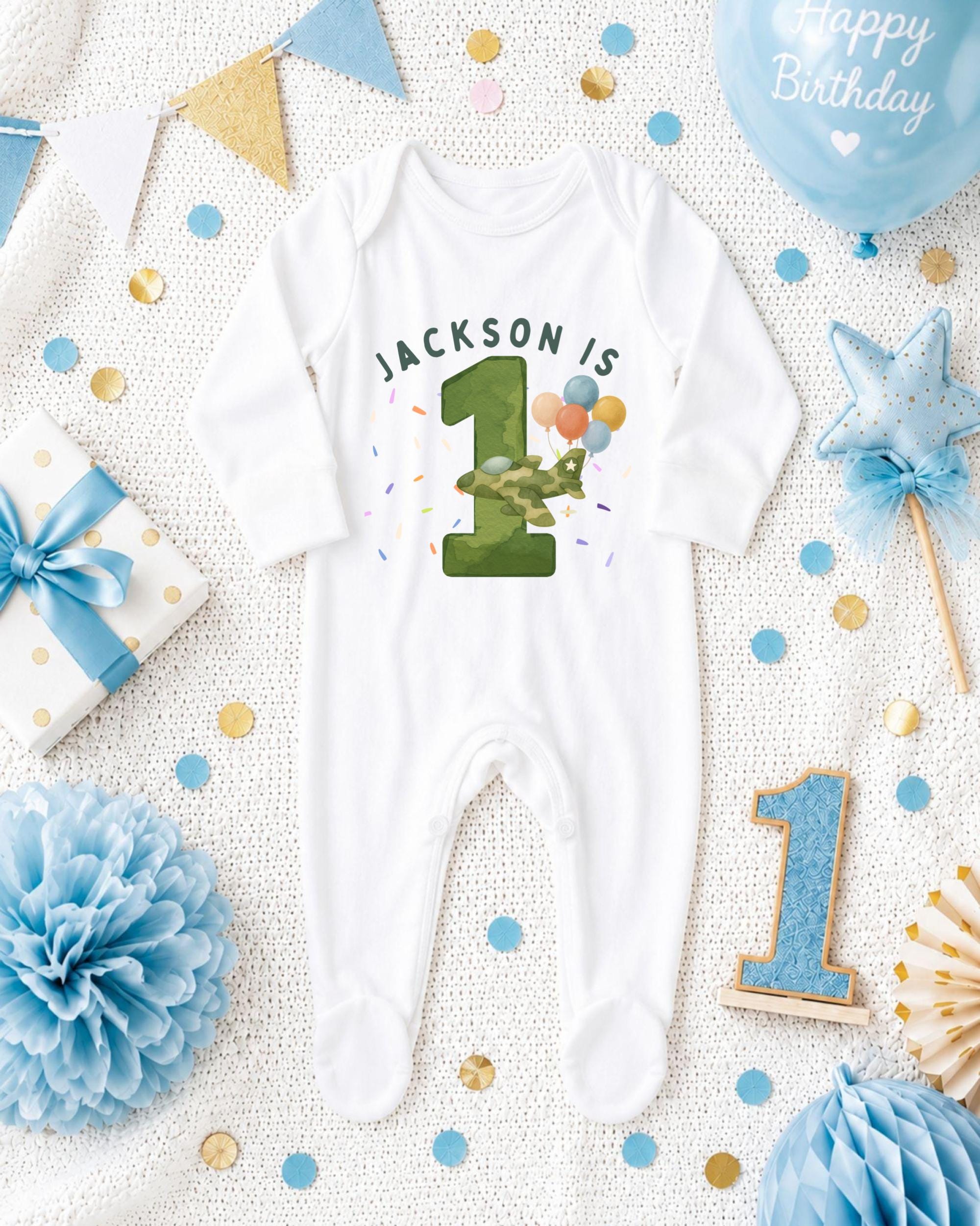 A white onesie with the number 1 and the text "JACKSON IS 1" printed on it, surrounded by birthday decorations such as a blue balloon, a star, and confetti.