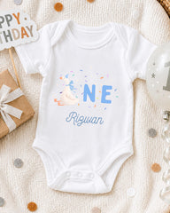 A white onesie with the text "Rizwan" printed on it, along with a birthday sign and a balloon, all placed on a white blanket with confetti.
