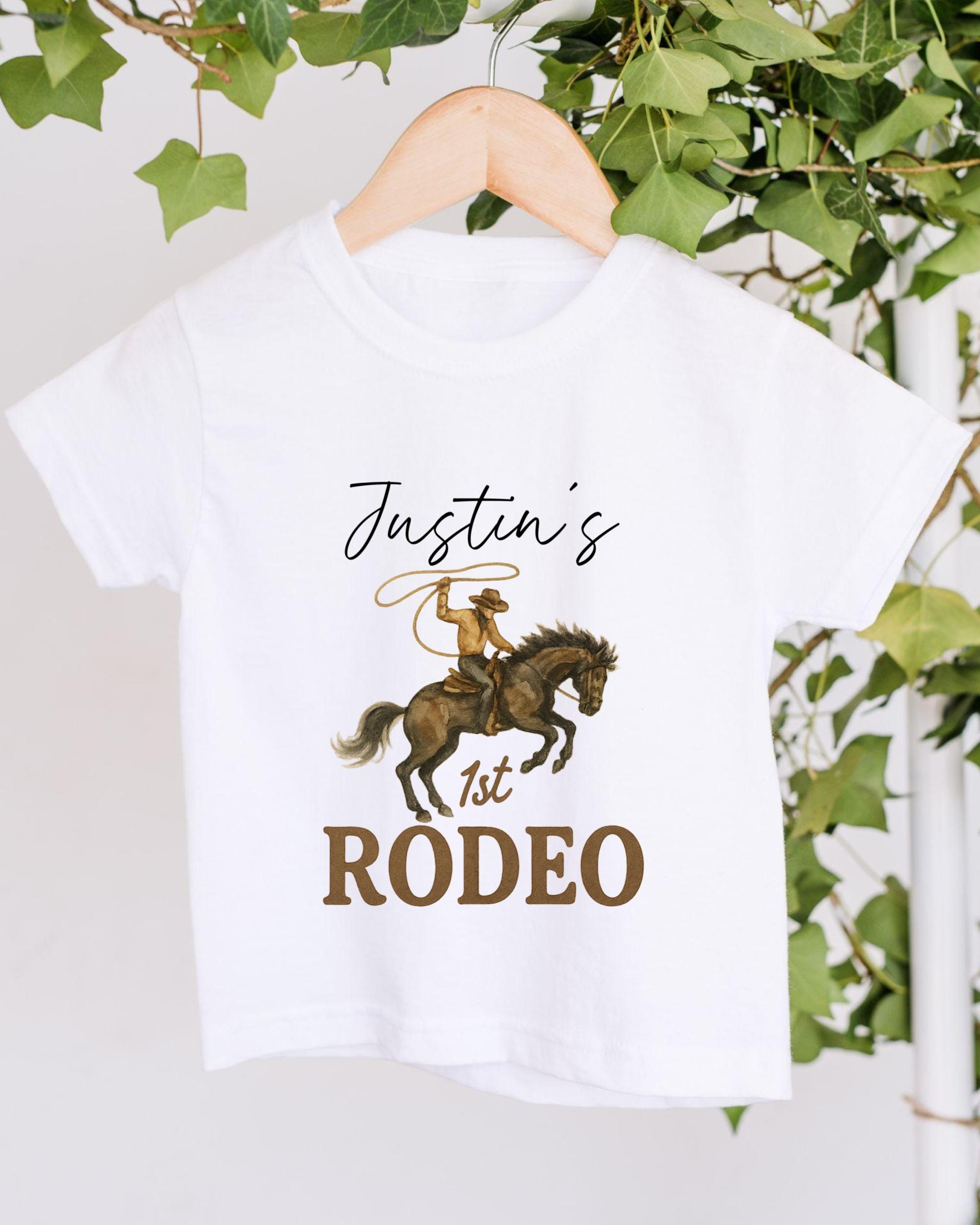 A white t-shirt with a cowboy graphic and text printed on it, hanging on a wooden hanger.