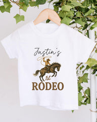 A white t-shirt with a cowboy graphic and text printed on it, hanging on a wooden hanger.