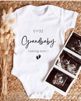 A white onesie with the text "First Grandbaby Coming Soon!" printed on it, along with a pair of footprints. The onesie is placed next to a set of three black and white images, which appear to be ultrasound images of a baby.