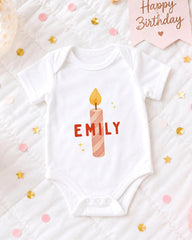 A white onesie with a candle design and the name "EMILY" printed on it, surrounded by confetti and a birthday card.