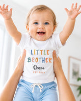A smiling baby wearing a white shirt with the text "Little Brother Oscar Est 2x" is being held up by someone's arms.