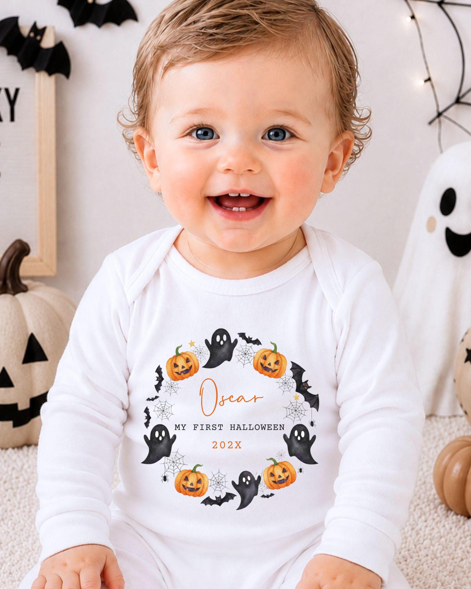 A smiling baby wearing a white onesie with a Halloween-themed design, including bats, pumpkins, and a ghost.