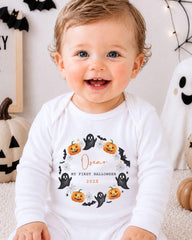 A smiling baby wearing a white onesie with a Halloween-themed design, including bats, pumpkins, and a ghost.