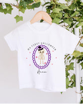 A white t-shirt with a Halloween-themed graphic design hanging on a wooden hanger. The design features a skeleton wearing a witch's hat and surrounded by hearts, with the text "My first Halloween Anna" displayed above the image.