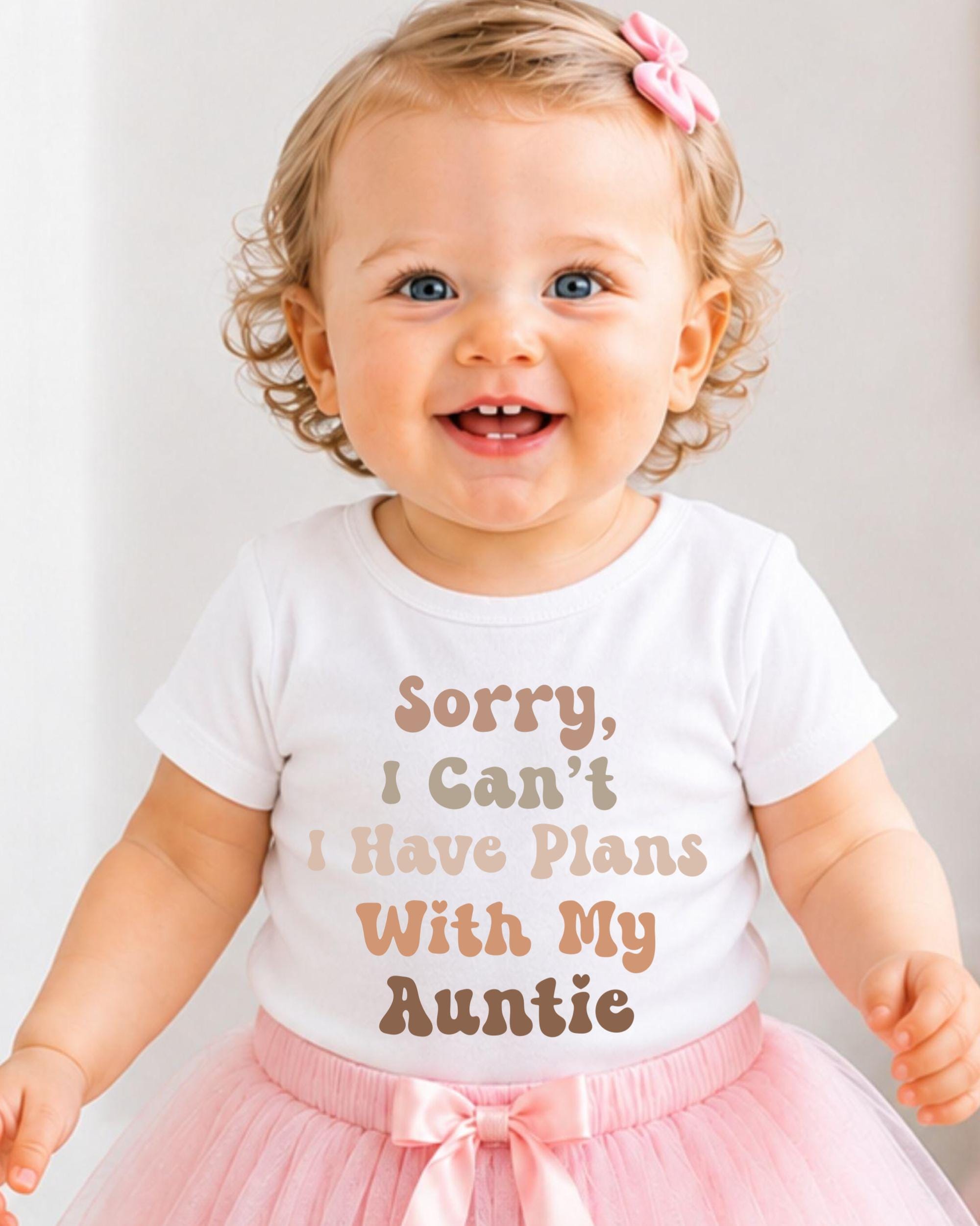 A smiling baby girl wearing a white t-shirt with text and a pink bow in her hair.