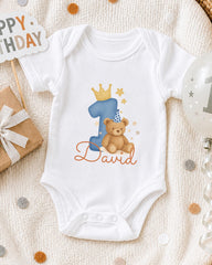 A white onesie with a blue number one and a teddy bear, along with a "Happy Birthday David" sign, is displayed on a bed with confetti.
