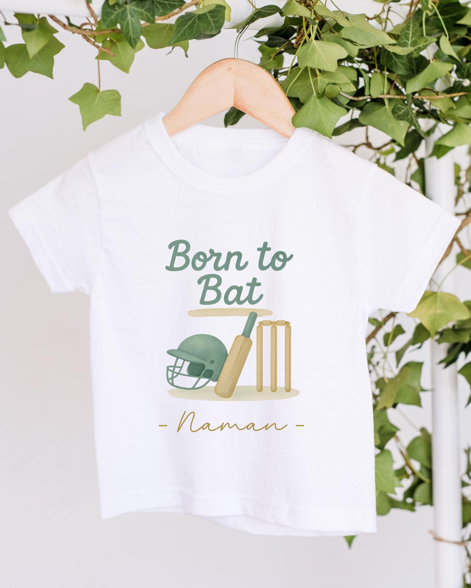 A white t-shirt with a graphic design featuring a cricket helmet, bat, and wickets, along with the text "Born to Bat" and "Naman" printed on it.