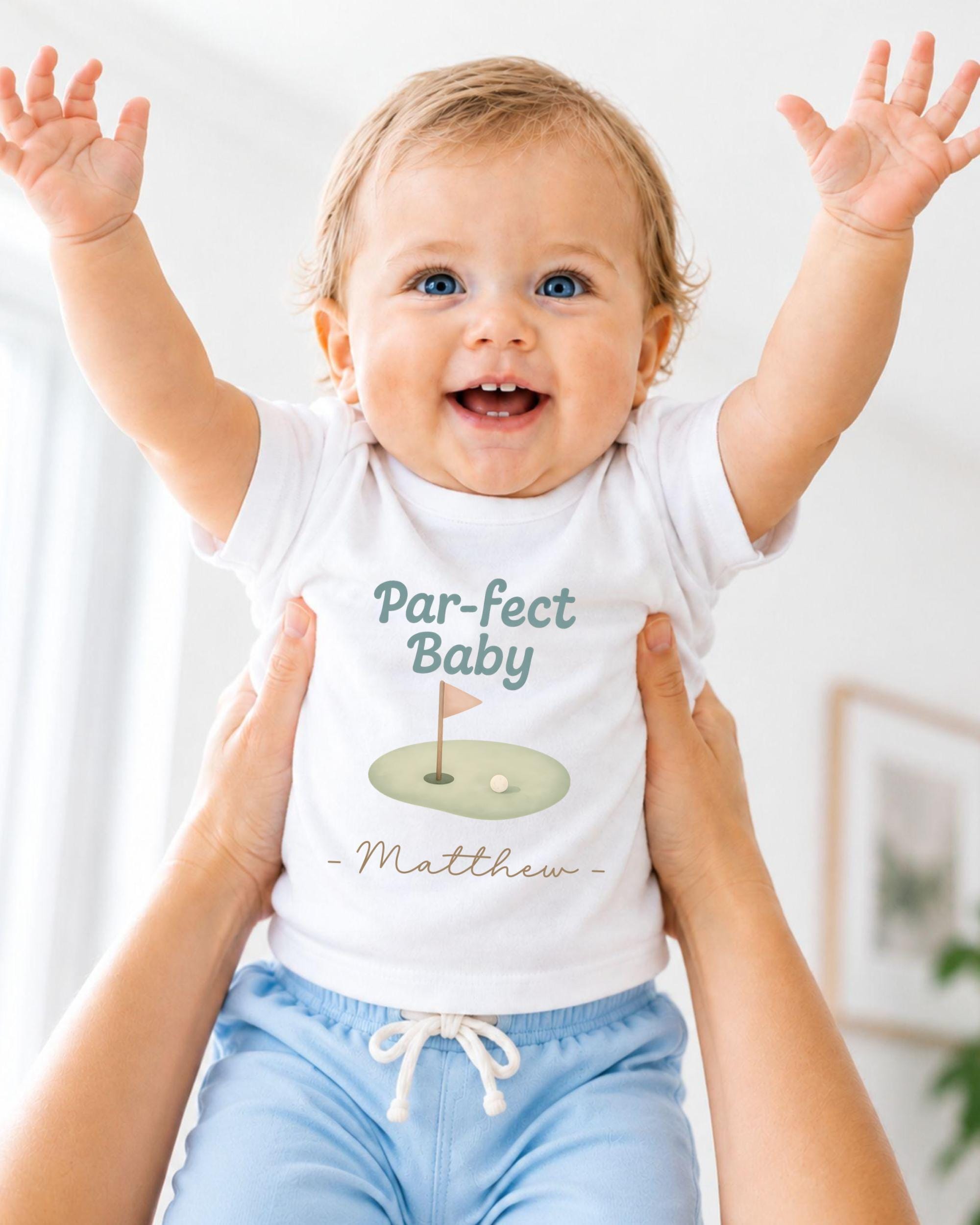 A smiling baby wearing a white t-shirt with a golf hole graphic and the text "Par-fect Baby Matthew" is being held up by someone's arms.