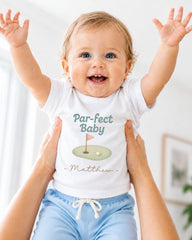 A smiling baby wearing a white t-shirt with a golf hole graphic and the text "Par-fect Baby Matthew" is being held up by someone's arms.