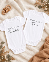 two white onesies with the text "Double the Trouble" printed on them, laid out on a white blanket with some floral decorations.
