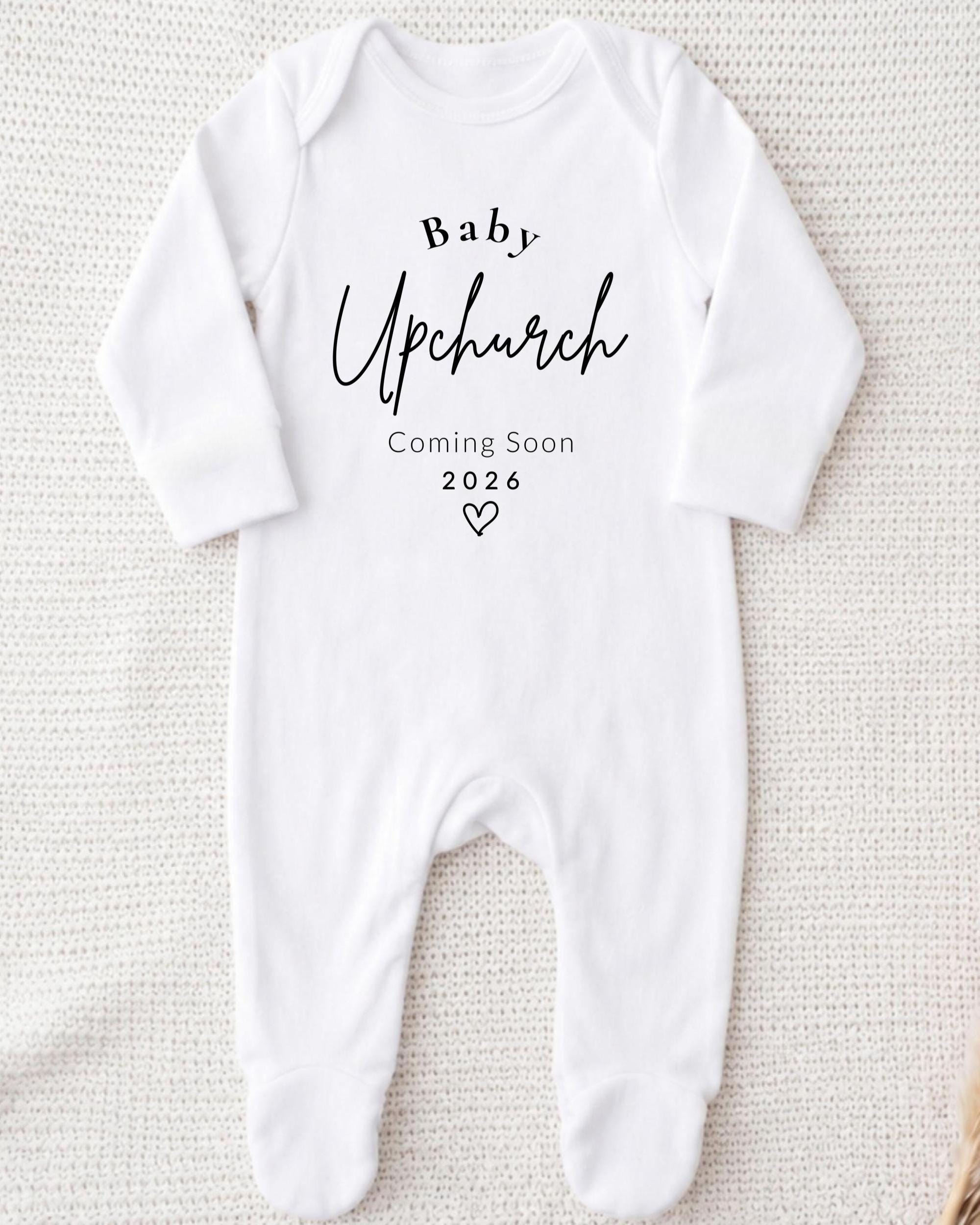 A white onesie with the text "Baby Upchurch" printed on it, along with the text "Coming Soon 2026" below it.