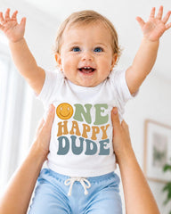 A smiling baby wearing a white shirt with the text "ONE HAPPY DUDE" is being held up by someone's arms.