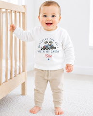 A smiling baby boy wearing a white sweatshirt with a racing theme and text, standing in a nursery setting next to a crib.