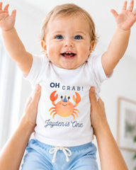 A smiling baby wearing a white t-shirt with a crab design and the text "OH Crab! Jayden is One" is being held up by someone's arms.