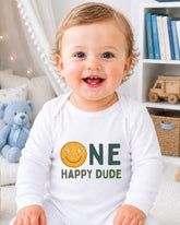 A smiling baby wearing a white onesie with a smiley face and the text "ONE HAPPY DUDE" printed on it.