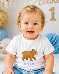 A smiling baby boy wearing a white t-shirt with a bear graphic and the name 'WILSON' printed on it, sitting in front of a birthday party backdrop.