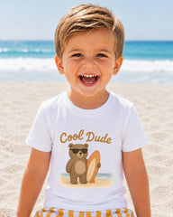 A young boy with a big smile on his face, wearing a white t-shirt with a cartoon bear and the text "Cool Dude" printed on it, standing on a sandy beach with the ocean in the background.